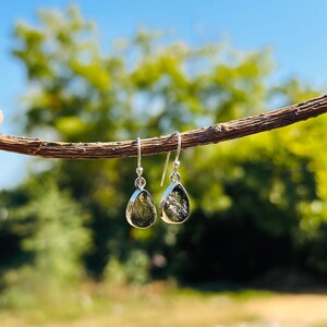 May include: A pair of silver dangle earrings with green teardrop-shaped stones. The earrings are hanging from a branch.