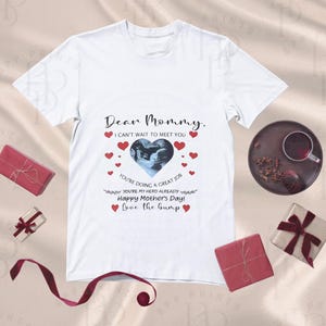 May include: White t-shirt with the text "Dear Mommy, I can't wait to meet you" and a heart-shaped sonogram image. Red hearts surround the image. Several wrapped gifts and a cup of tea are also visible.