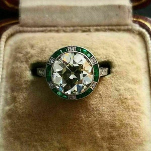 Pode incluir: Um anel de noivado de diamante branco redondo com um halo de pedras verde esmeralda. O anel é engastado em uma banda prateada e é apresentado em uma caixa de anel de veludo.