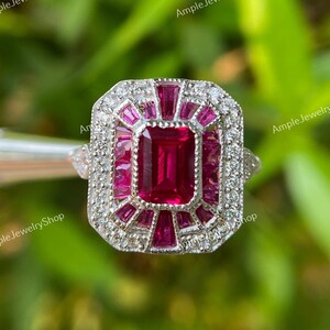 Könnte beinhalten: Ein silberner Ring mit einem rechteckigen, tiefroten Mittelstein, umgeben von kleineren roten und klaren Steinen. Der Ring hat ein geometrisches Design und ist vor einem verschwommenen grünen Hintergrund platziert. Der Ring ist von Ample Jewelry Shop.