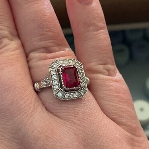 May include: A silver ring with a large, red, emerald-cut gemstone surrounded by smaller diamonds.