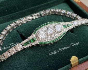 Op de afbeelding: Een zilveren armband met heldere en groene stenen ligt in een houten sieradendoos met een groene fluwelen voering. De armband heeft vierkant geslepen stenen en een ovaal middenontwerp. De doos is van hout.