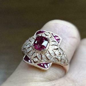 Könnte beinhalten: Ein goldener Ring mit einem großen, quadratischen Rubin, der von kleineren Diamanten umgeben ist. Der Ring hat ein filigranes Design und ist in eine Weißgoldfassung gefasst.