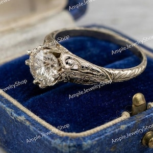 May include: A silver-toned ring featuring a round, faceted clear stone set in a prong setting. The band and the gallery of the ring are decorated with intricate, engraved scrollwork patterns throughout the metal surface.