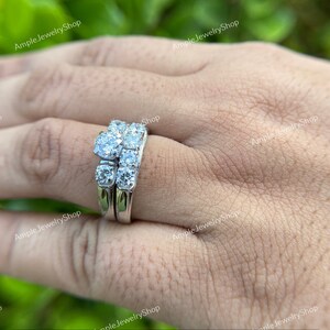May include: A silver wedding ring set with a large round center stone and smaller stones on the band. The ring is on a person's finger.