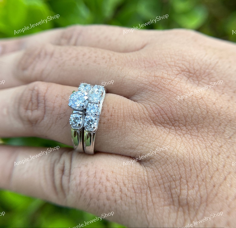 May include: A silver wedding ring set with a large round center stone and smaller stones on the band. The ring is on a person's finger.