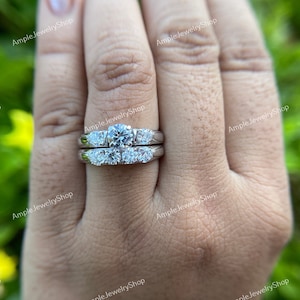 May include: A silver wedding ring set with a large round center stone and smaller stones on the band. The ring is on a person's finger.