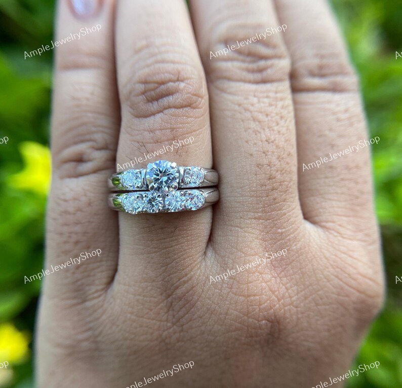 May include: A silver wedding ring set with a large round center stone and smaller stones on the band. The ring is on a person's finger.