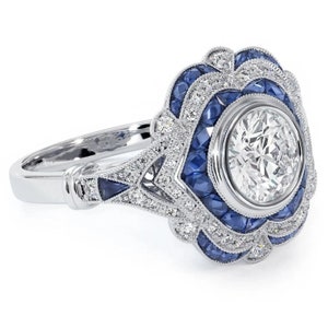 Peut inclure: Une bague en argent avec une grande pierre ronde centrale entourée d'un halo de petits saphirs bleus et de diamants. La bague présente un délicat motif floral aux détails complexes.