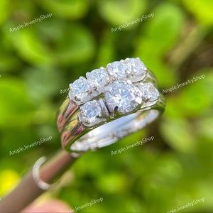 May include: A silver wedding ring set with a large round center stone and smaller stones on the band. The ring is set on a green background.