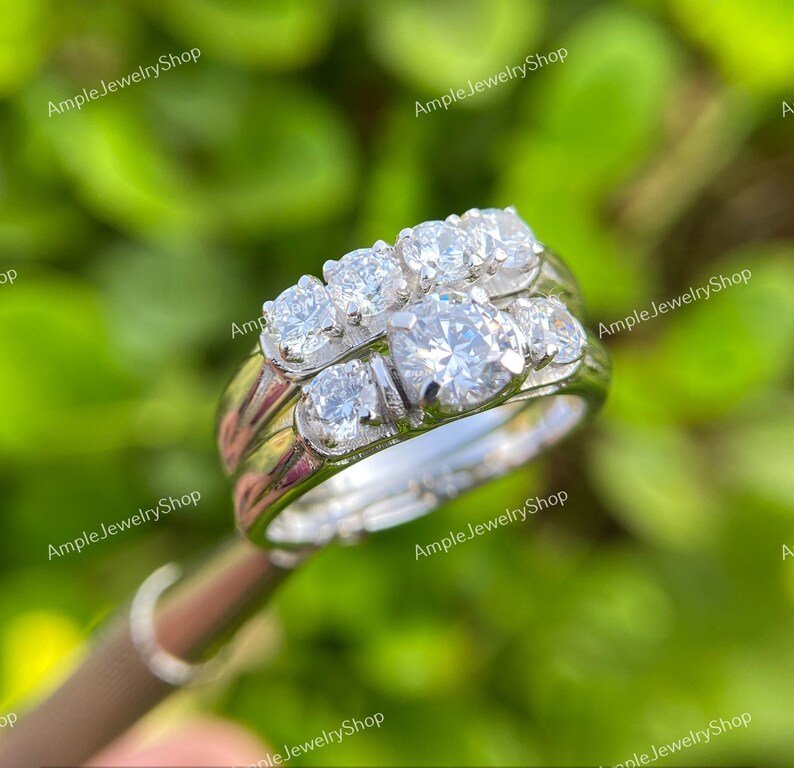 May include: A silver wedding ring set with a large round center stone and smaller stones on the band. The ring is set on a green background.