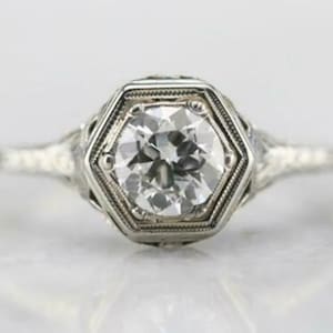 May include: A close-up of a silver ring with a large, clear, round-cut diamond set in a hexagonal frame. The ring has intricate filigree detailing on the band and around the diamond setting, creating a vintage aesthetic.