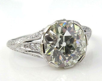 May include: A vintage engagement ring with a large, round, brilliant-cut diamond set in a platinum band. The diamond is surrounded by smaller diamonds and the band is decorated with intricate filigree.