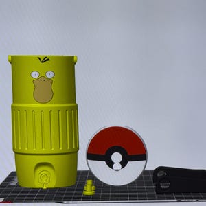May include: A yellow Psyduck-themed water bottle with a cartoon face, a red and white Pok&eacute; Ball, and a black accessory. The bottle has a cylindrical shape with vertical grooves and a handle. The Pok&eacute; Ball is a circular design.