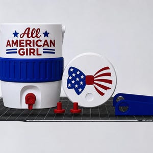 May include: A white and blue cooler with the text "All AMERICAN GIRL" in red and blue. The cooler has a blue band and a red spigot. A white lid with a bow tie design in red and blue, two red stoppers, and a blue handle are also present.
