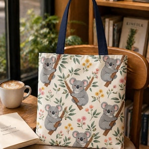May include: Cream-colored tote bag featuring a repeating pattern of gray koalas perched on tree branches with green leaves and colorful flowers. The bag has navy blue handles and is sitting on a wooden chair.