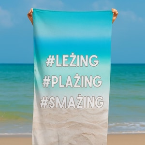 May include: Beach towel with a gradient blue and white design, featuring the hashtags #LEZING, #PLAZING, and #SMAZING in white. The towel is held up against a beach and ocean backdrop.