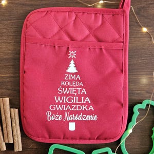 May include: A red quilted oven mitt with a white Christmas tree design and Polish text. Beside it are green cookie cutters and cinnamon sticks, all arranged on a wooden surface. A string of fairy lights adds a festive touch.