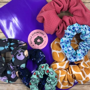 May include: A set of five colorful hair scrunchies in a purple bag. The scrunchies are made of fabric and have different patterns, including a galaxy print, a mermaid scale print, a honeycomb print, a green print with hearts, and a solid red print. The bag has a sticker that says "Battle Scrunchies Handmade Like us on Facebook".