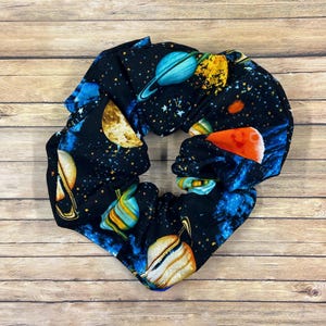 May include: A black scrunchie with a colorful space-themed pattern. The fabric features planets, stars, and nebulas in shades of blue, orange, yellow, and white. The scrunchie is made of a soft, gathered fabric.