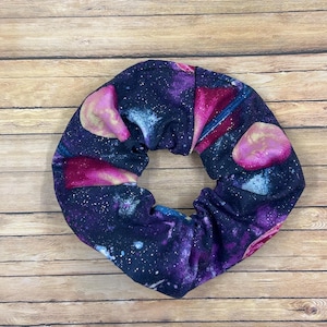 May include: A scrunchie with a galaxy print featuring shades of purple, pink, and blue, with white star-like details. The fabric is gathered, creating a circular shape. The scrunchie is set against a wooden background.