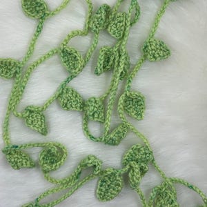 May include: A handmade crochet garland featuring green leaves and vines. The leaves are a light green color with darker green accents, and the vines are a similar shade. The garland is designed to be flexible and can be used for various decorative purposes.
