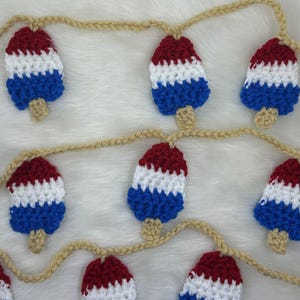 May include: A crochet garland featuring red, white, and blue popsicle shapes. The popsicles are strung together with a beige yarn, creating a festive and patriotic decoration. The garland is perfect for adding a touch of summer fun to any space.