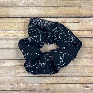 May include: A black fabric hair scrunchie featuring a white celestial print with various constellations, stars, and zodiac signs including Pisces and Libra. The design includes thin white lines connecting star clusters and celestial motifs.