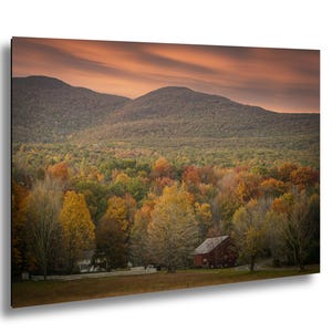 May include: A landscape photograph of a rural scene during autumn. The image features a vibrant display of fall foliage in shades of yellow, orange, and red, with a red barn and white house in the distance. Mountains and an orange sky complete the scene.
