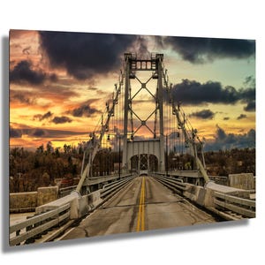 May include: A suspension bridge with a steel structure and a road leading to the center. The bridge is surrounded by trees and a cloudy sky with a dramatic sunset in the background.