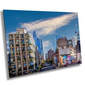 May include: A view of a city street in New York City with tall buildings, a blue sky with white clouds, and a street with cars and people walking.