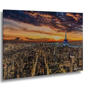May include: A rectangular wall art print featuring an aerial view of the New York City skyline at sunset. The Empire State Building is illuminated with blue lights, surrounded by a dense grid of city lights under a vibrant orange and purple sky.