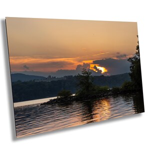 May include: A scenic sunset over a lake with a small island in the foreground. The sky is a vibrant orange and pink, with clouds reflecting in the water. The sun is setting behind the distant hills.