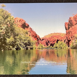 May include: A scenic view of a red rock gorge with a calm body of water in the foreground. The water reflects the sky and the red rock cliffs. The text "Outback 2 Ocean Photography" and "Adel's Grove Gorge, QLD" is visible at the bottom of the image.