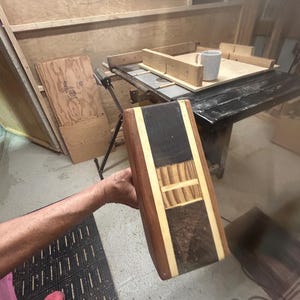 May include: A rectangular wooden object with a multi-toned design. The object features sections of dark brown, light brown, and black wood, with lighter wood accents. The item is held by a person, suggesting it is a handmade craft or woodworking project.