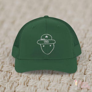 May include: A green trucker hat with a white embroidered outline of a leprechaun face. The hat features a mesh back and a curved brim. The leprechaun's hat has a shamrock on it. The hat is a St. Patrick's Day themed accessory.