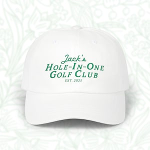 May include: White baseball cap with a curved peak. The cap displays the text "Jack's Hole-In-One Golf Club EST. 2025" in green lettering. A classic style hat, suitable for golf enthusiasts.