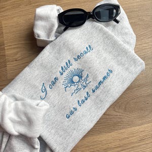 Op de afbeelding: Een lichtgrijs sweatshirt met blauw geborduurde tekst "I can still recall our last summer". Een zwarte zonnebril ligt op de gevouwen sweatshirt. Een witte scrunchie is ook zichtbaar.