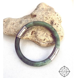 Könnte beinhalten: Ein jadegrünes Armreif-Armband mit silberfarbenen Metallakzenten. Das runde Armband liegt auf einem hellen Stein. Die Metallverschlüsse sind mit einer filigranen Kette verbunden. Der Hintergrund ist eine weiße Oberfläche.