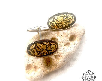 May include: A pair of oval cufflinks with a black background and gold floral design. The cufflinks are connected by a silver bar and rest on a light-colored, textured stone. The design features intricate details.