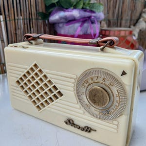 Op de afbeelding: Crèmekleurige vintage radio met een ruitvormig luidsprekerrooster en een afstemknop. De radio heeft een handvat met een roze band en de merknaam "Sonett" staat op de voorkant. Een klassiek retro-ontwerp.