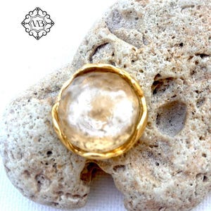 Könnte beinhalten: Ein Ring mit einem runden, durchscheinenden, champagnerfarbenen Stein mit texturierter, gehämmerter Oberfläche, gefasst in eine polierte, goldfarbene Metallfassung mit gewelltem Rand. Ein kleines schwarzes Logo mit den Buchstaben VB ist oben links zu sehen.
