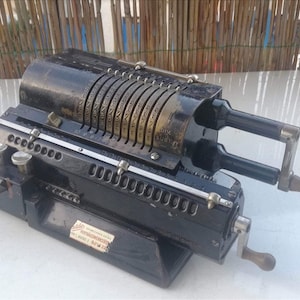 Op de afbeelding: Een zwarte vintage mechanische rekenmachine met meerdere wijzerplaten en twee handkrukken. De rekenmachine heeft een metalen plaat met de tekst "Mercedes Rechenmaschinenfabrik Berlin-Lichtenberg" en "Made in Germany".