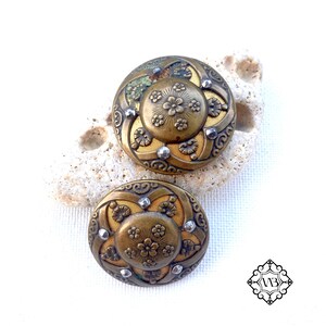 Könnte beinhalten: Zwei antike Messingknöpfe mit floralen Designs und silberfarbenen Akzenten. Die Knöpfe haben eine goldfarbene Mitte und sind vor einem weißen Hintergrund platziert. Die Knöpfe sind wahrscheinlich zum Nähen oder Basteln.