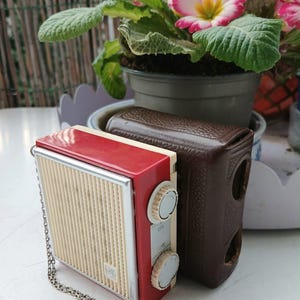 Könnte beinhalten: Ein rotes und cremefarbenes Vintage-Radio mit einer silbernen Kette. Das Radio hat einen cremefarbenen Lautsprechergrill und zwei weiße Bedienknöpfe. Eine braune Ledertasche befindet sich neben dem Radio. Eine Topfpflanze mit rosa Blüten ist im Hintergrund.