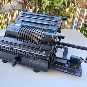 Può includere: Una calcolatrice meccanica vintage nera con una manovella e una serie di quadranti numerati. La calcolatrice ha il testo "Brunsviga Maschinen-Aktien-Gesellschaft" e "Grimme, Natalie & Co., Ltd." stampato sul lato.
