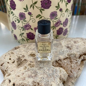 Può includere: Un piccolo flacone di profumo Chanel N°5 Eau de Toilette in vetro trasparente. Il flacone ha un tappo nero e un'etichetta con le parole "Eau de Toilette N°5 Chanel Paris".