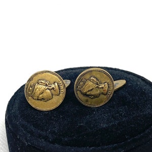 May include: A pair of gold-toned cufflinks featuring a profile of a person's head with the word "SOLIVA" inscribed on the edge.