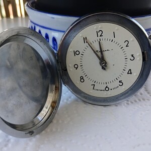Può includere: Un orologio da tasca argentato con un quadrante bianco e numeri neri. L'orologio è aperto e il retro è visibile. Il quadrante dell'orologio dice "СССР" e "Москвa".