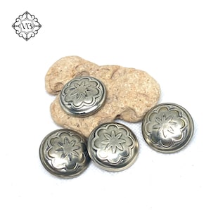 Op de afbeelding: Vier zilverkleurige, ronde metalen knopen met een bloemmotief worden getoond op een lichtbruine steen. De knopen hebben een verhoogd, gedetailleerd patroon. Een logo staat in de linkerbovenhoek.